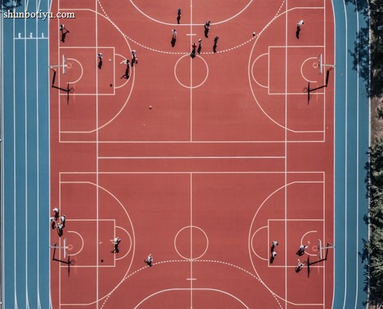 An aerial view of a large, vibrant sports complex featuring a red basketball court with multiple hoops and a surrounding blue running track. Several people are scattered across the area, engaged in various activities.