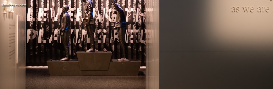 A dimly lit display features statues of athletes standing on a podium. They are positioned in front of a backdrop with the words 'LEVELING THE PLAYING FIELD' in bold letters. On the wall to the right, a softly lit quote is partially visible. The overall ambiance suggests a museum or exhibition setting.