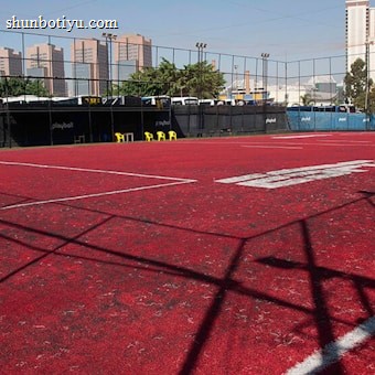 A sports court with a red playing surface and white lines, surrounded by a black mesh fence. In the background, tall buildings are visible, along with some trees and a few yellow chairs positioned near the fence.